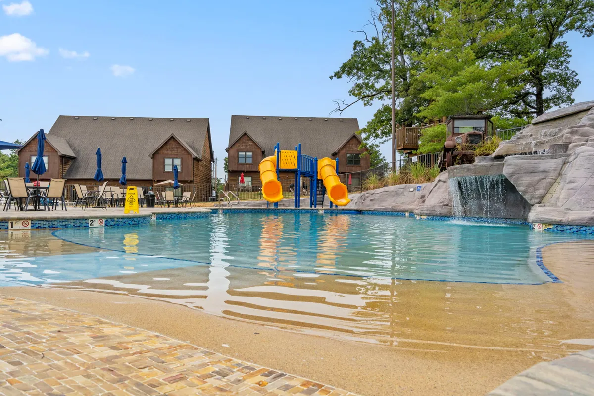 A vibrant outdoor scene featuring the Branson Shores Resort's swimming pool, complete with a water slide, waterfall, lounge chairs, and surrounding cabins. Several groups of people are enjoying the pool and relaxing in the sun, while children play in the water and adults gather on the poolside deck.