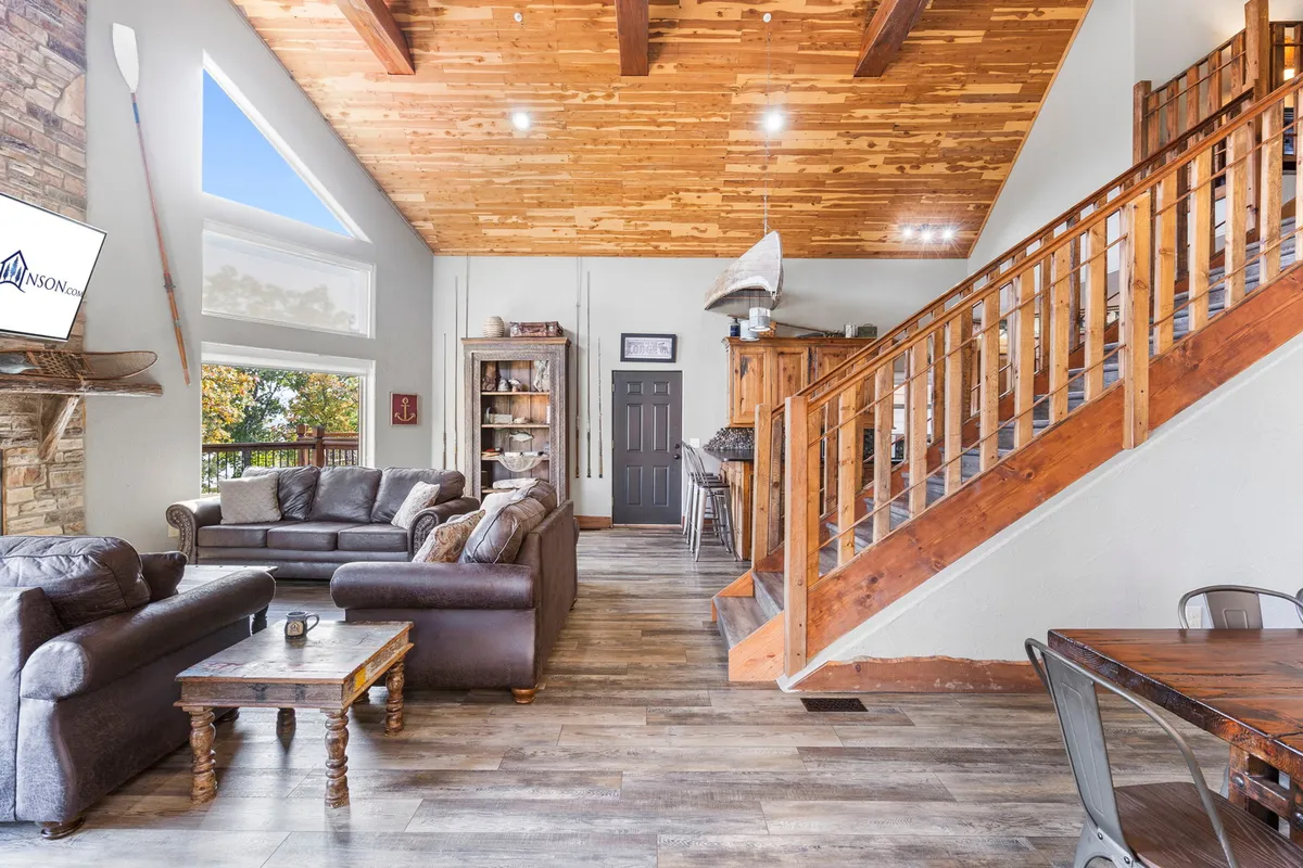 A welcoming open-plan space that includes a cozy sofa set, visible wooden beams, a staircase, television, and a dining area just beyond. The layout seamlessly connects living, dining, and kitchen zones, ideal for group gatherings and easy flow between spaces.