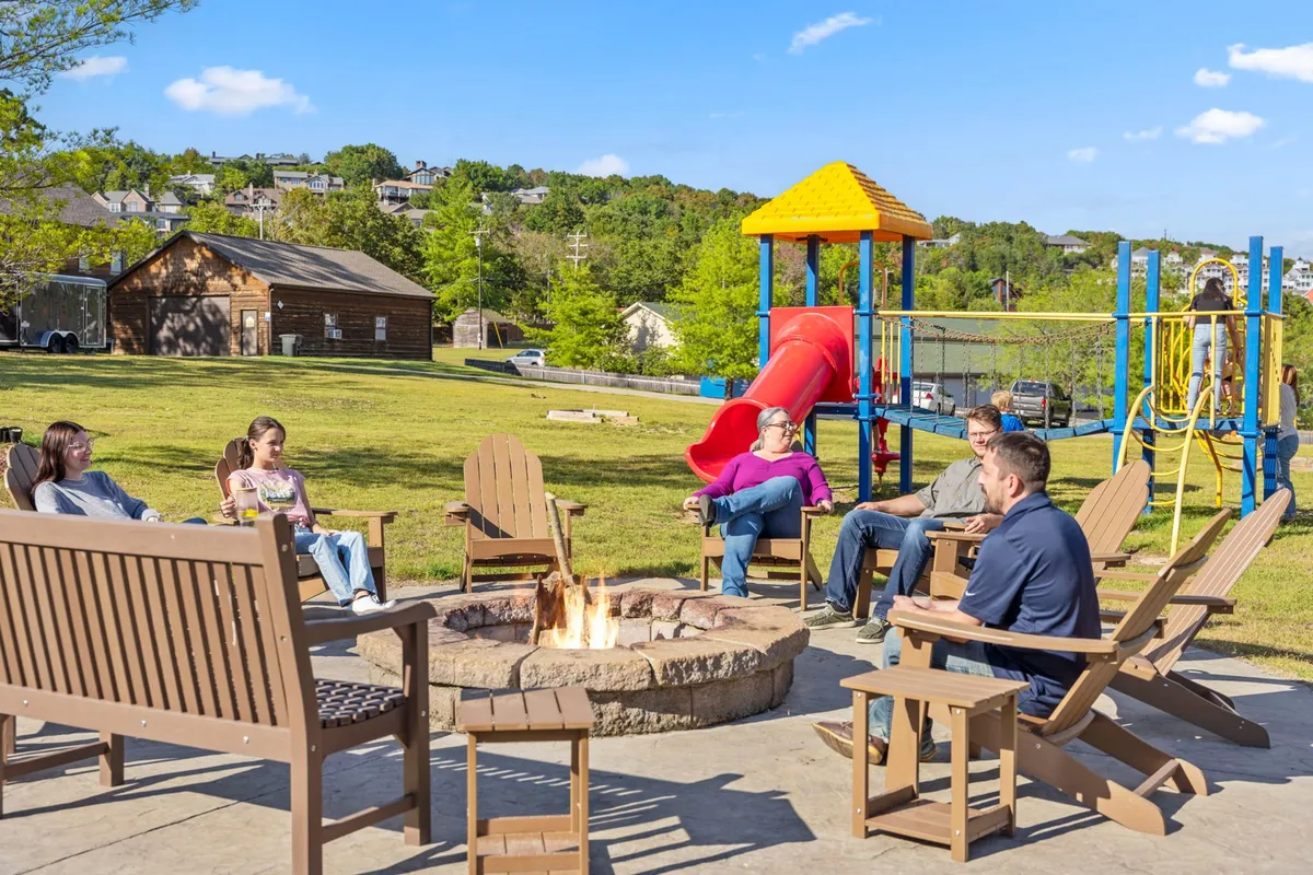 An exterior shot focused on a cozy fire pit area featuring a seating circle, playground equipment, and people gathered around the fire. Children are seen playing in the background while adults enjoy each other’s company, all set against a backdrop of a glowing sunset.