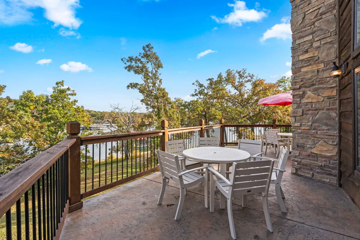 An exterior patio scene with a table and chairs overlooking the water, surrounded by natural wood railings and lush greenery. The setting is serene, perfect for outdoor dining, gathering with friends, and soaking in the sunset over the water.