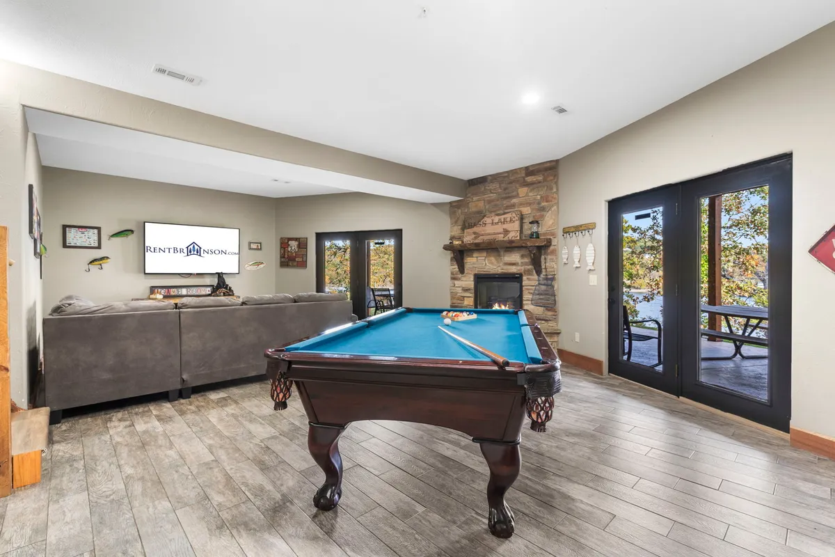 A spacious and inviting recreation room featuring a pool table in the foreground, a cozy stone fireplace, large windows bringing in natural light, and a comfortable sofa arranged for group gatherings and TV watching.