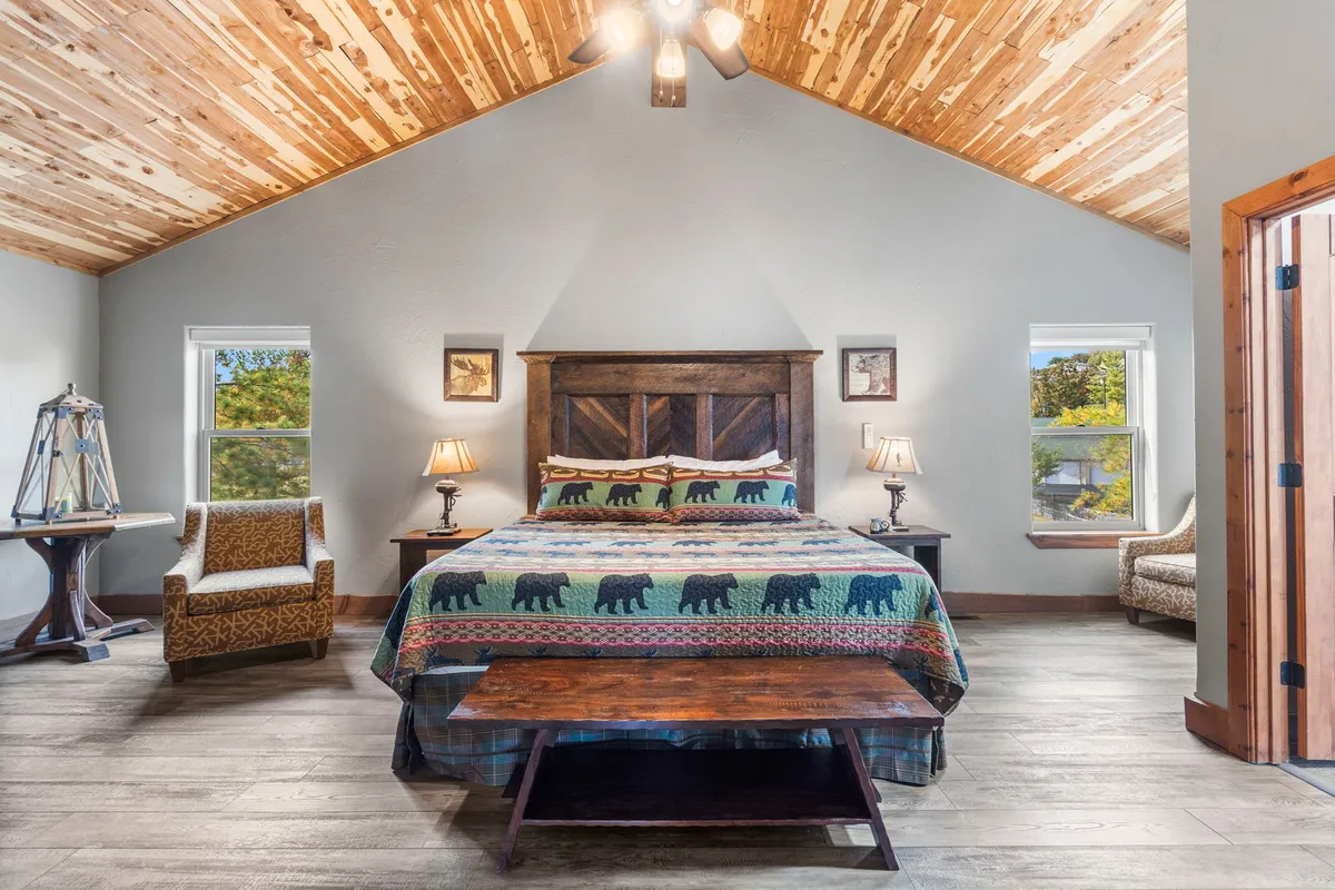 A comfortable king bedroom with rustic touches, featuring a bed with bear-themed bedding, wooden ceiling, two bedside lamps, a seating area with a chair, and a wooden coffee table. The ambiance is warm and inviting, ideal for restful nights.