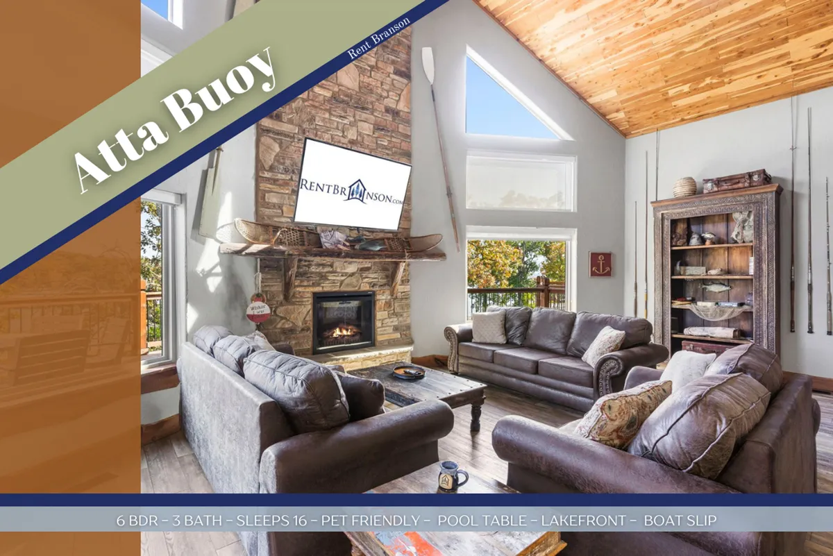 A spacious living room featuring a cozy stone fireplace, large windows overlooking the lake, plush seating, and a pool table. The real-wood ceiling and warm lighting enhance the room’s inviting, rustic feel. Antique canoes may be visible as unique decor touches, while the windows frame the beauty of Table Rock Lake.