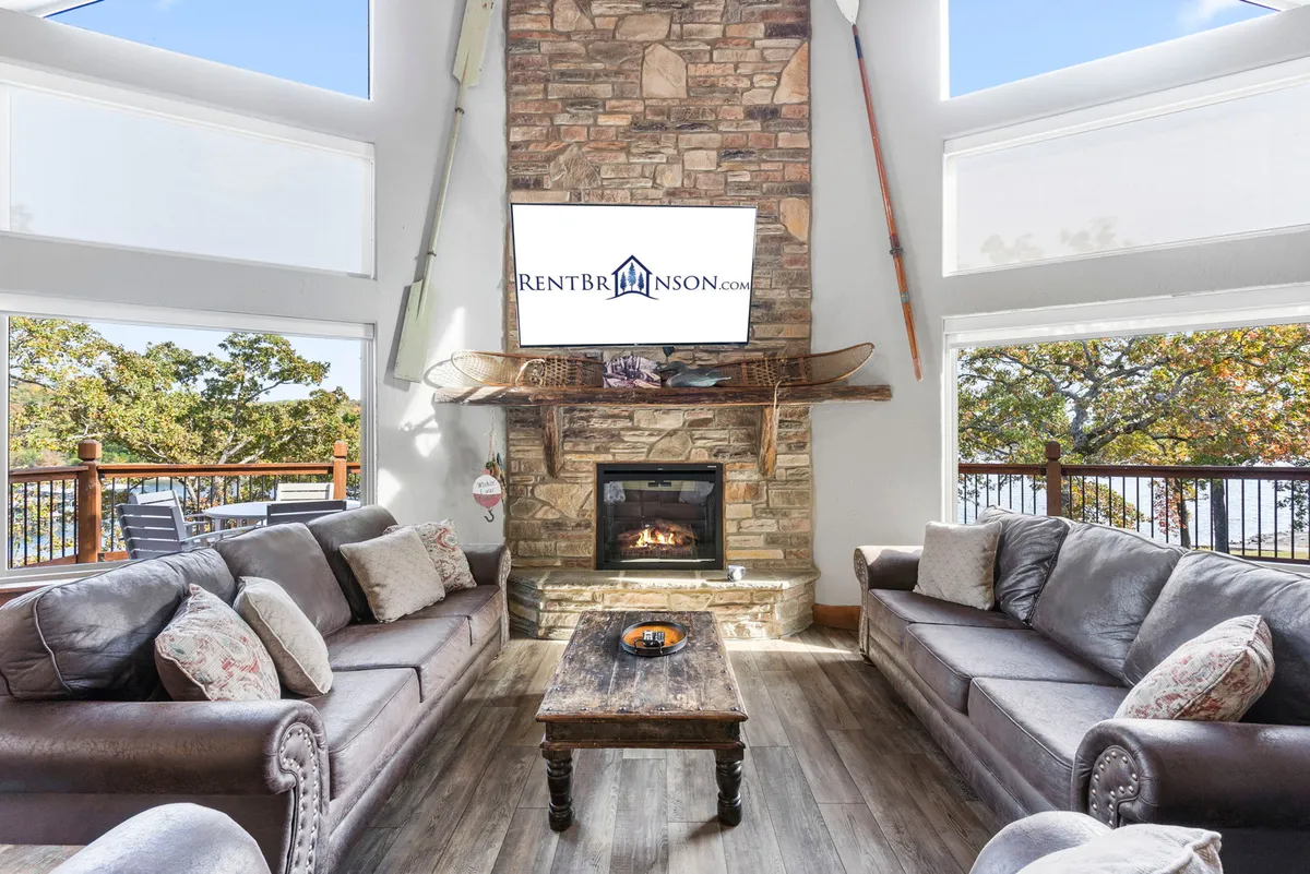 A cozy interior living space centers on a stone fireplace, large windows revealing scenic outdoor views, and comfortable seating. The room is bathed in soft light, inviting guests to relax and enjoy the warmth of the fire during colder months.