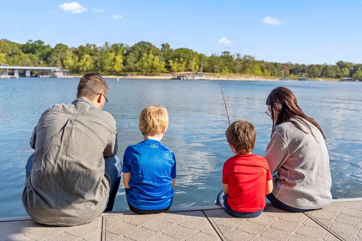 A family sits together by the calm water’s edge, fishing rods in hand, surrounded by nature. The scene exudes joy and serenity, capturing the fun and relaxation of a lakeside fishing excursion and quality time outdoors.