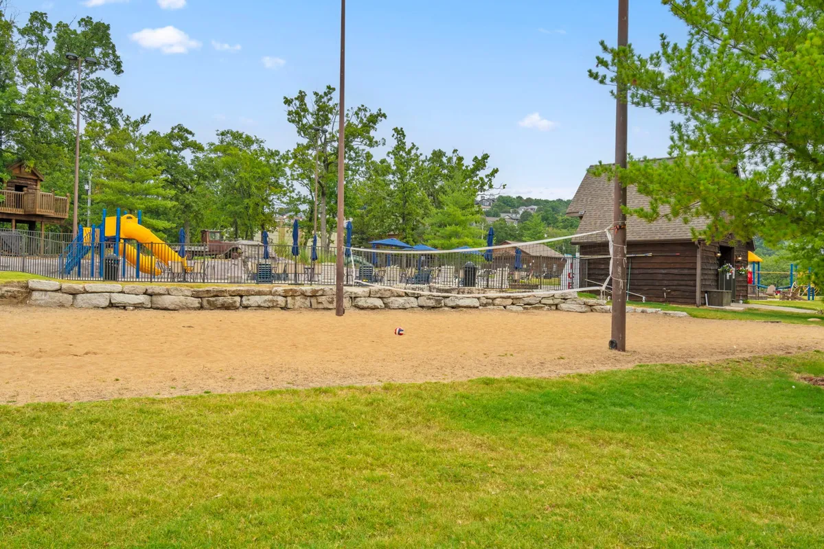 A dynamic exterior shot of the sand volleyball court adjacent to the playground, with families picnicking, children playing, and lush trees all around. The image suggests an active, welcoming environment where guests can easily transition from sports to relaxation.
