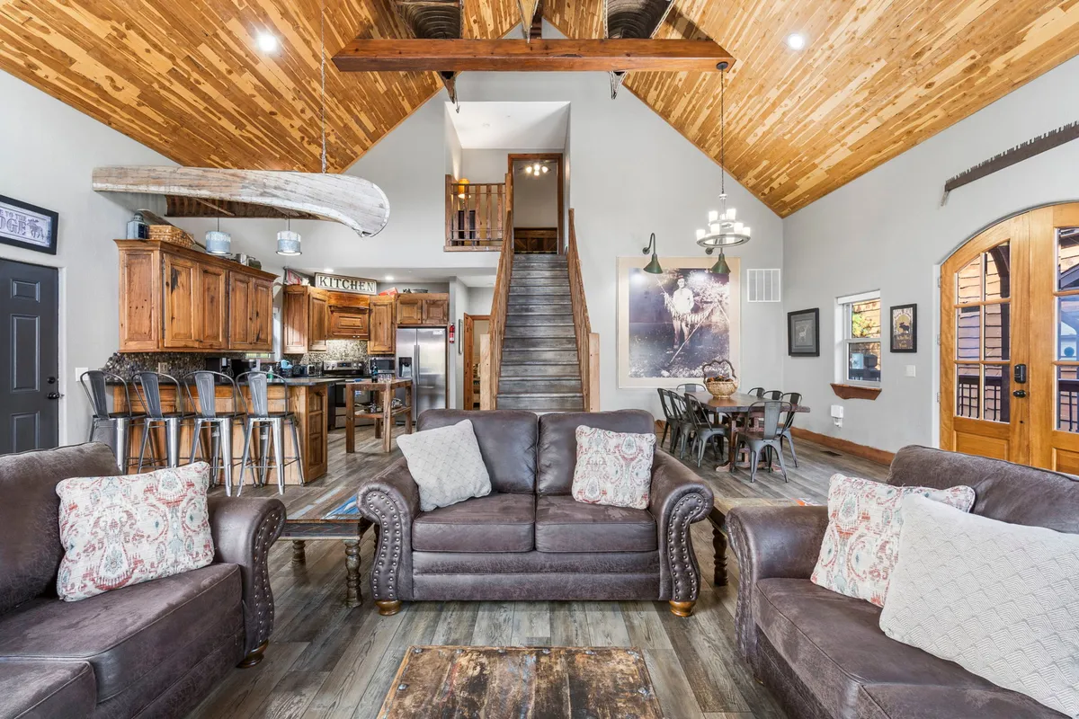 The image showcases a spacious, warmly lit open-concept living area featuring plush sofa seating beneath rustic wooden beams, a large dining table perfect for shared meals, a modern kitchen, and a welcoming staircase—all designed for family gatherings and relaxation.