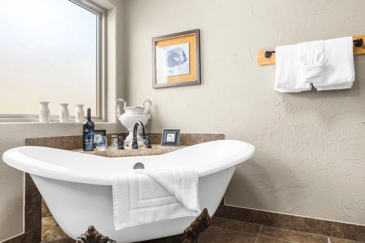 A tranquil master suite bathroom scene with a freestanding claw-foot tub, decorative vases, framed artwork, and plush towels. The soft lighting and elegant finishes evoke relaxation and luxury.