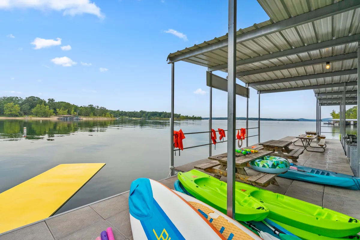 Kayaks and paddleboards rest on a floating dock at the water’s edge, with picnic tables and calm water stretching into the distance. The setting is tranquil and adventure-ready, inviting guests to launch watercraft or enjoy a peaceful moment by the lake.